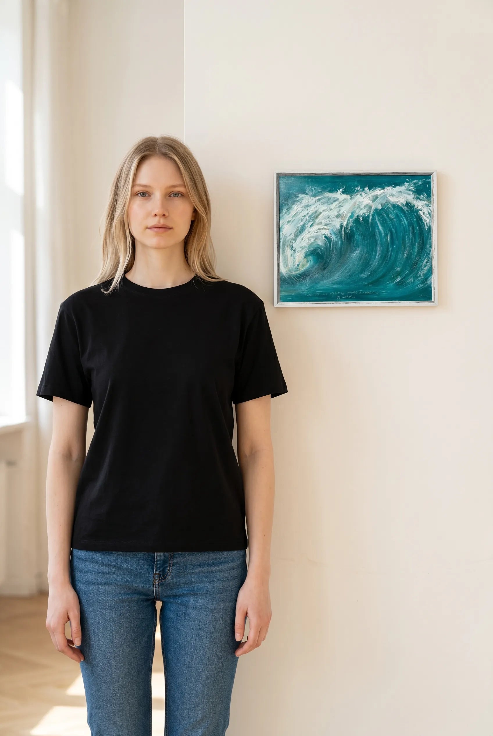 Person wearing a black t-shirt and blue jeans standing in a room with a painting of a wave by Charlotte Gibbs on the wall.