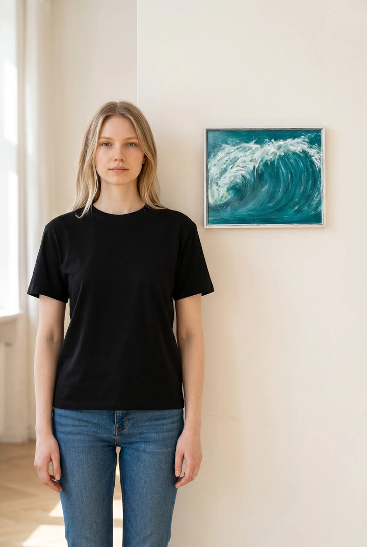 Person wearing a black t-shirt and blue jeans standing in a room with a painting of a wave by Charlotte Gibbs on the wall.