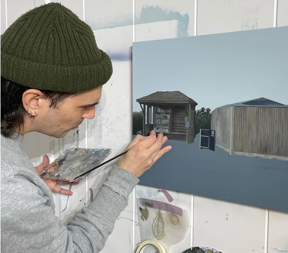 A person wearing an olive green beanie and gray sweatshirt paints a rustic outdoor scene on a canvas, holding a paint palette and brush in a studio setting.