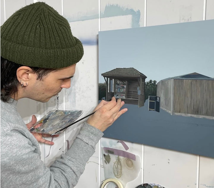 A person wearing an olive green beanie and gray sweatshirt paints a rustic outdoor scene on a canvas, holding a paint palette and brush in a studio setting.