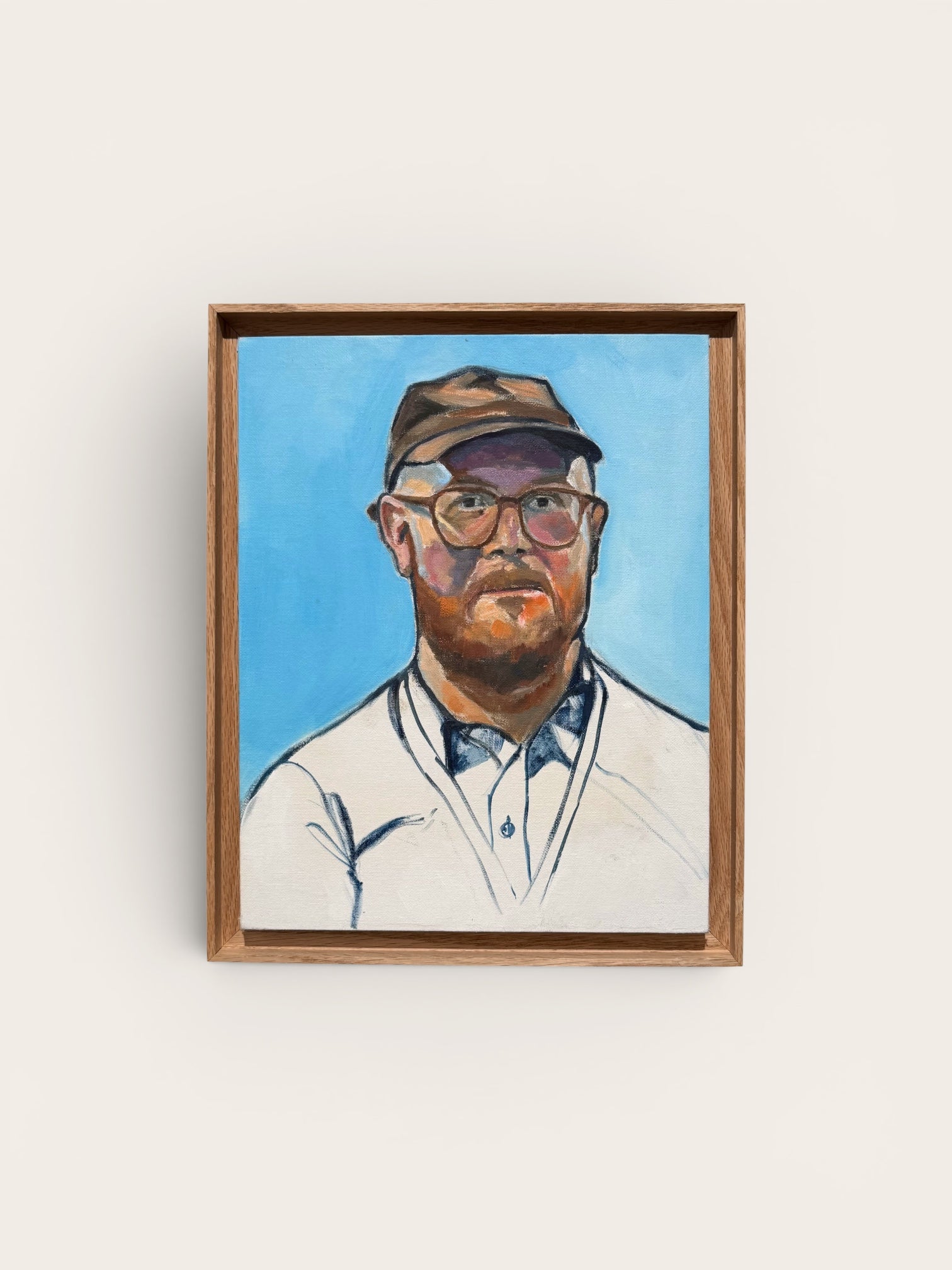 A framed portrait painting of a man with a beard, glasses, and a cap, set against a light blue background, hanging on a plain off-white wall.