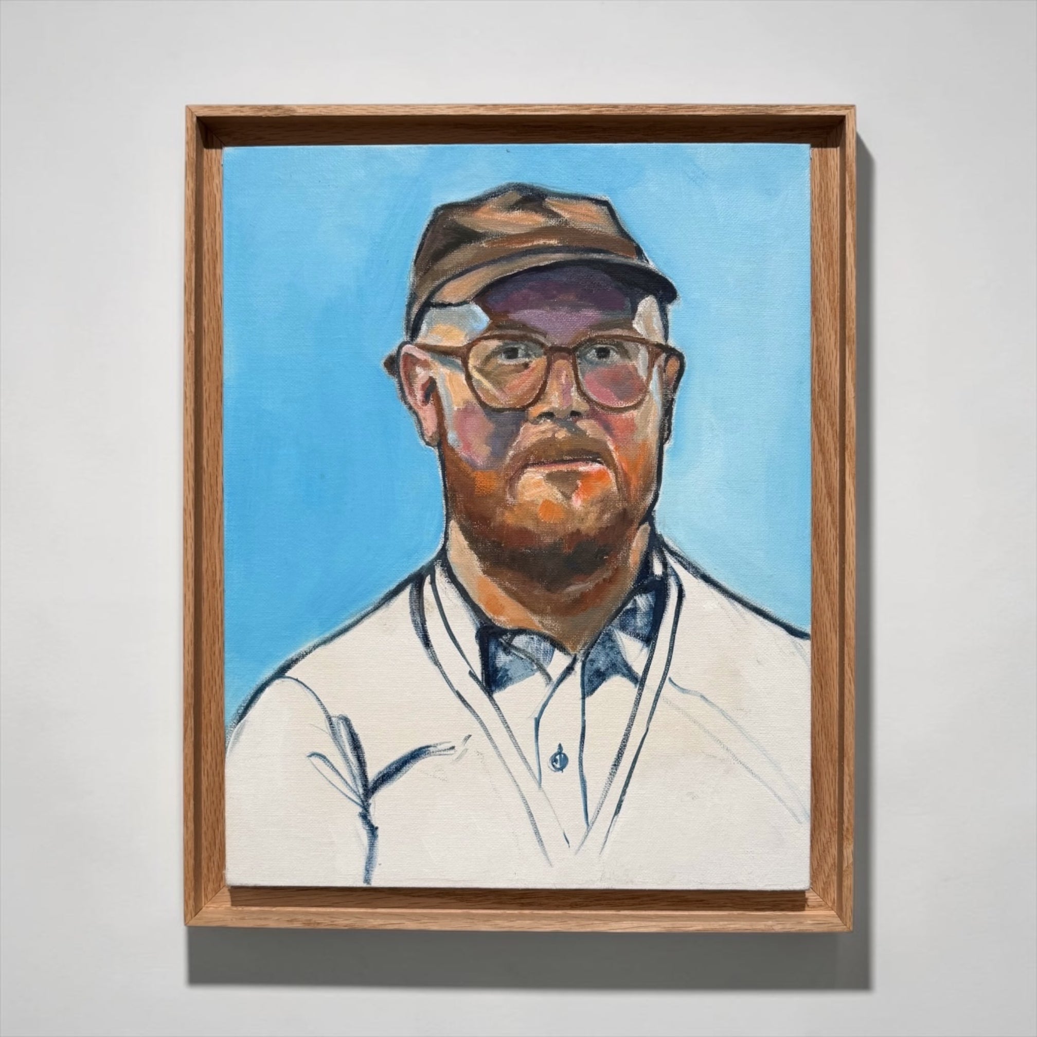 A framed portrait painting of a man with a beard, glasses, and a cap, set against a bright blue background, hanging on a light gray wall.