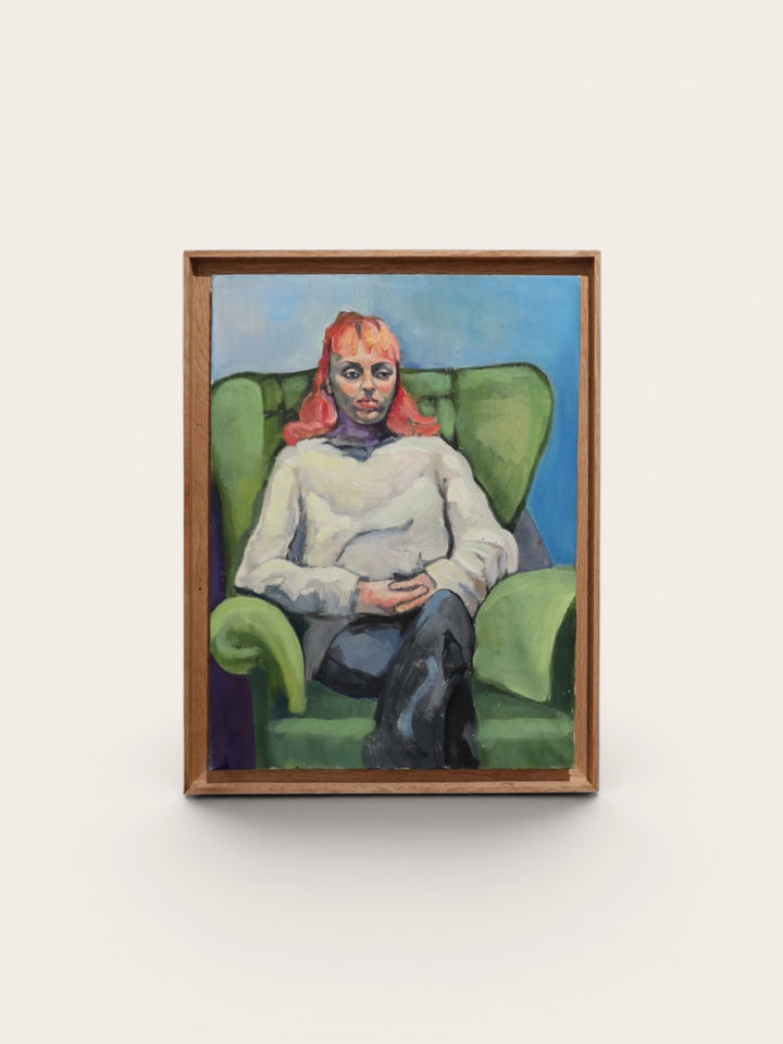 A framed portrait painting of a seated person with red hair, wearing a white top and dark pants, set against a green armchair and blue background, mounted on a light wall.