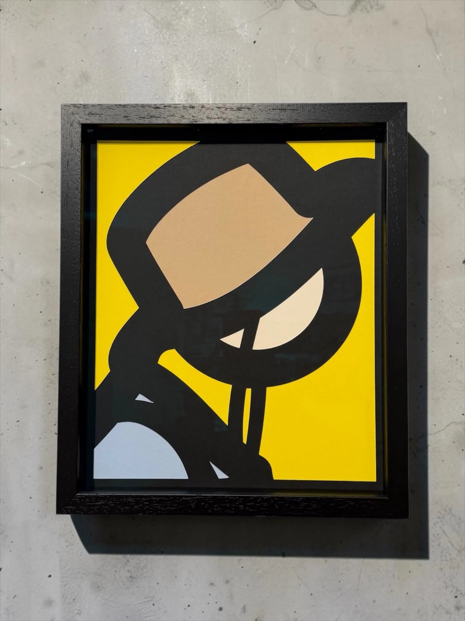 Framed artwork of a stylised paper cut face on a yellow background, leaning against a textured wall.