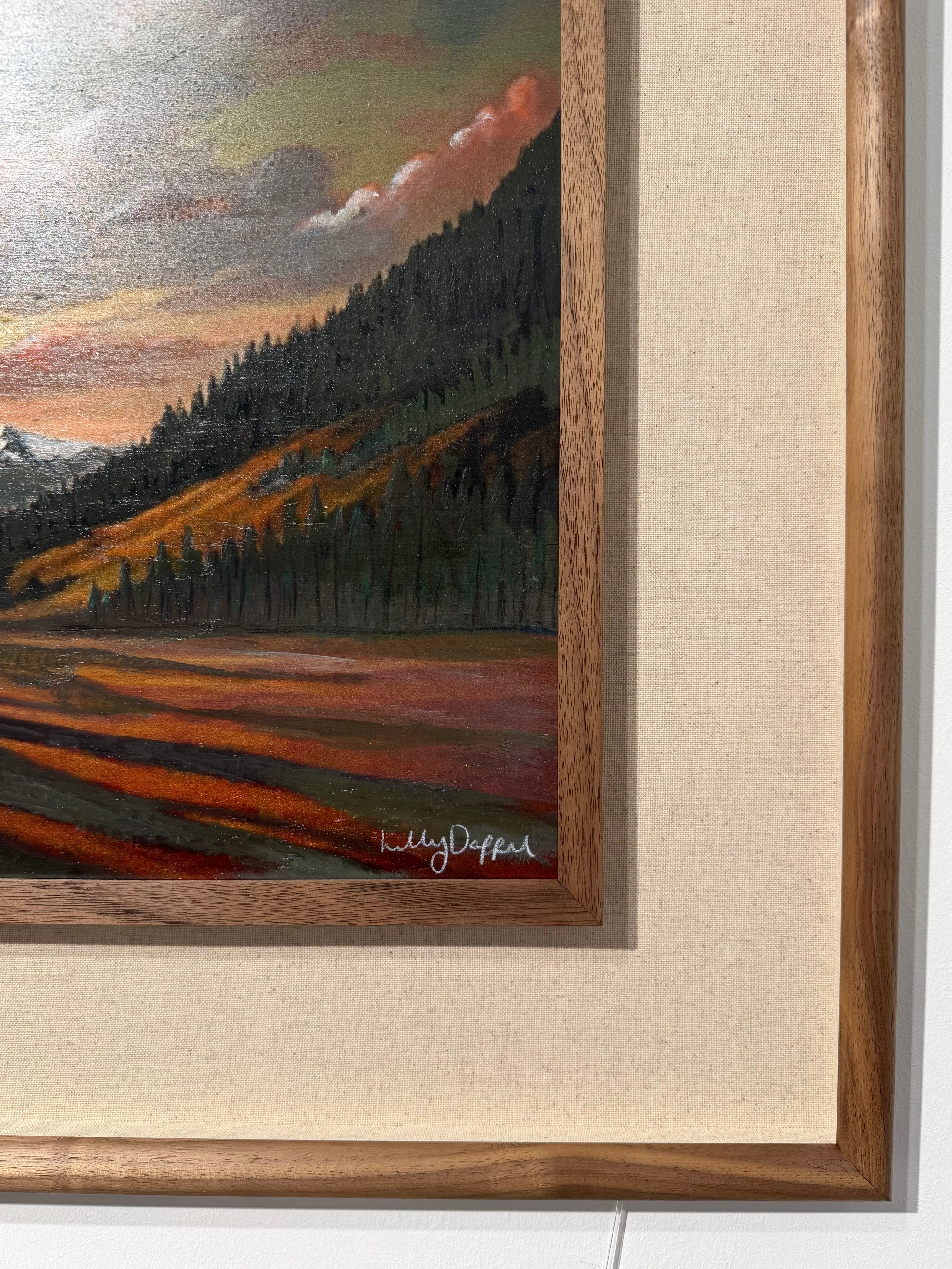 A framed landscape painting showing a forested mountain slope, fields, and a cloudy sky, with a visible artist signature near the lower right corner. The artwork is mounted with a wide cream mat and a medium brown wood frame.