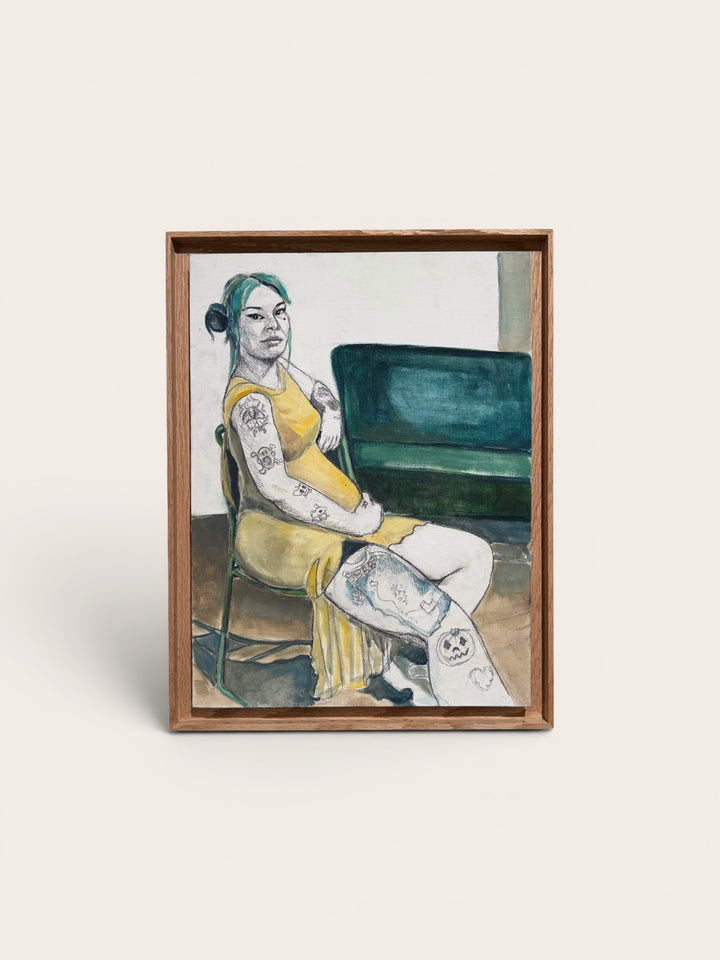 A framed artwork featuring a painted portrait of a tattooed woman in a yellow dress seated in front of a green chair, displayed against a light beige background.