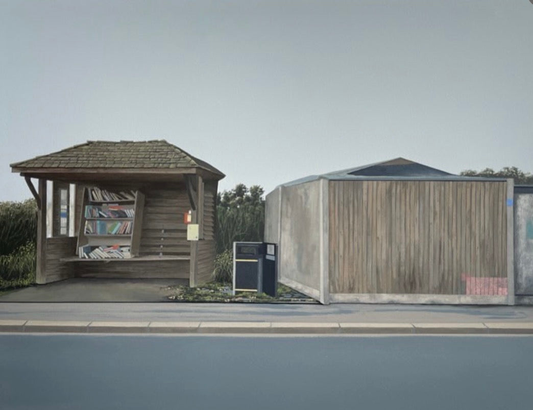 A wide outdoor scene showing a small wooden shelter with a bench and bookshelves beside a larger wooden structure, set near a road under a pale sky.