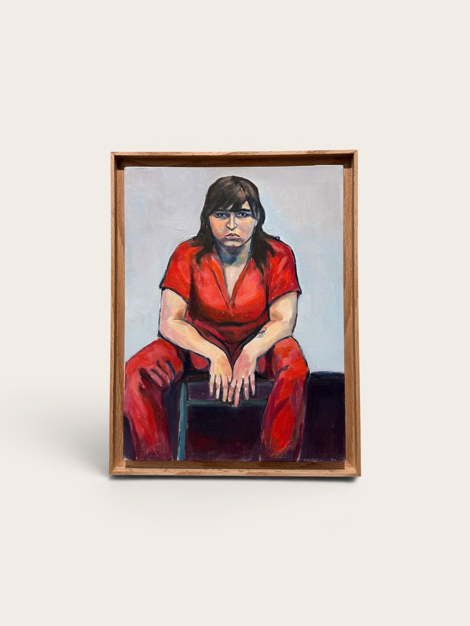 A framed portrait painting of a seated person in a bright red outfit against a pale background, shown on a light neutral backdrop. Ruth By Juan Manrique