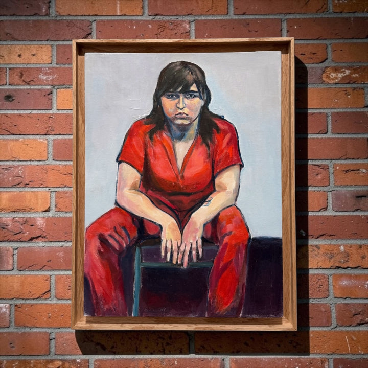 A framed portrait of Ruth by Juan Manrique painting of a seated woman in a bright red outfit, mounted on a red brick wall.