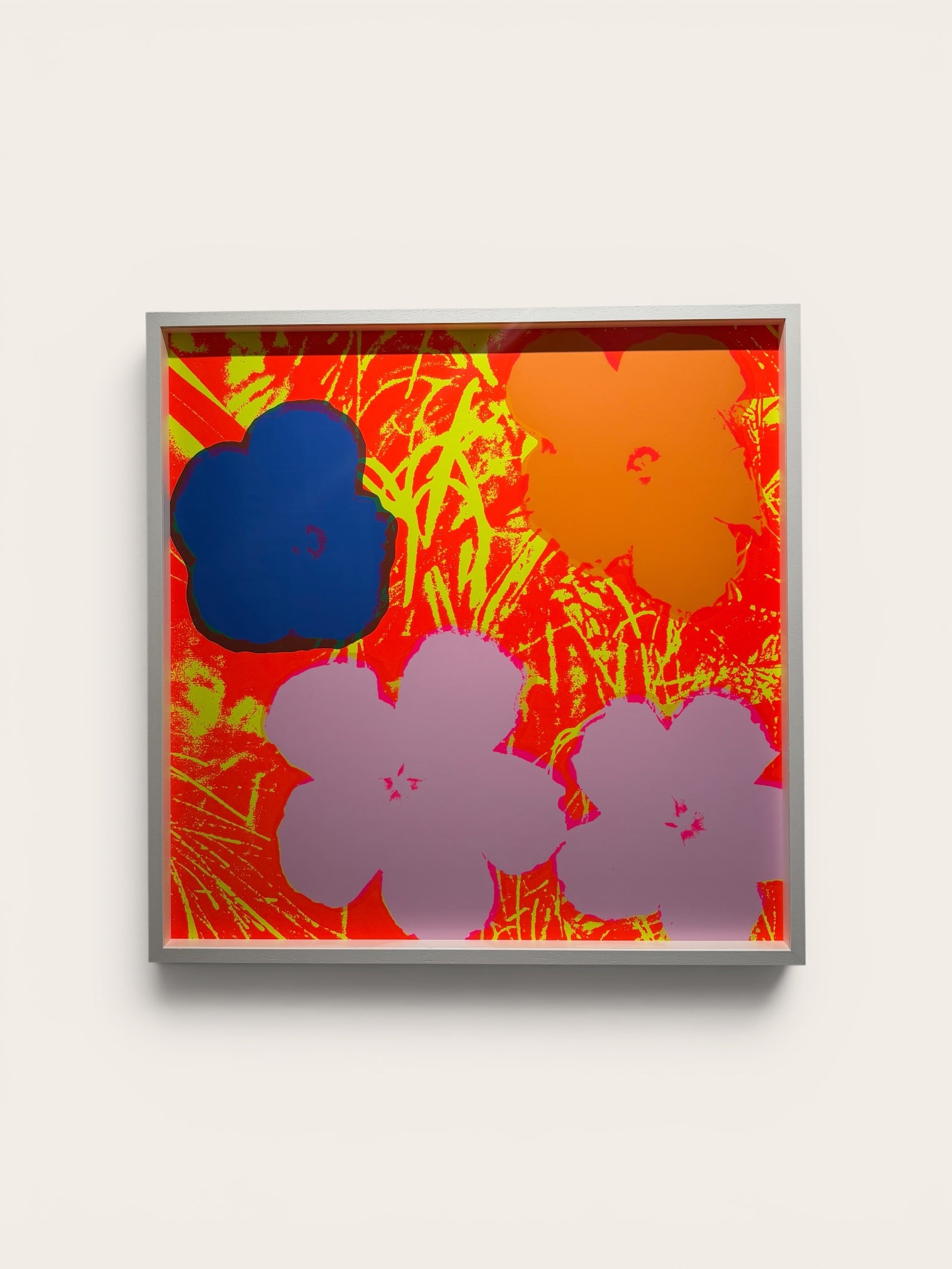 A square framed abstract art print featuring bold floral silhouettes in blue, orange, and lavender over a vibrant red and yellow background, mounted on a light beige wall.