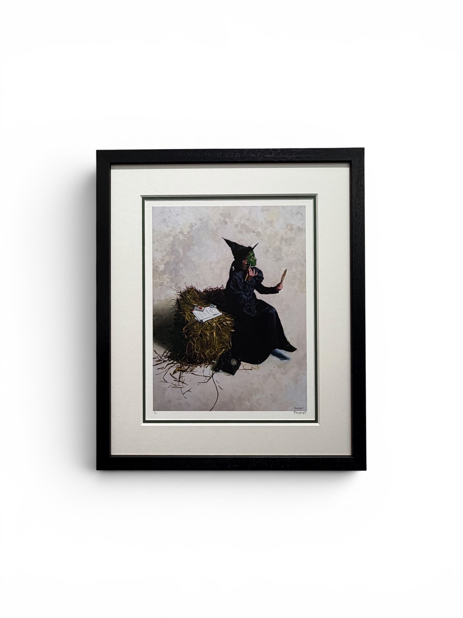 A framed art print featuring a dark illustration of a witch in a black hat and cloak beside a nest, set against a light background. The print is matted with a white border and enclosed in a black frame, shown against a plain white wall.
