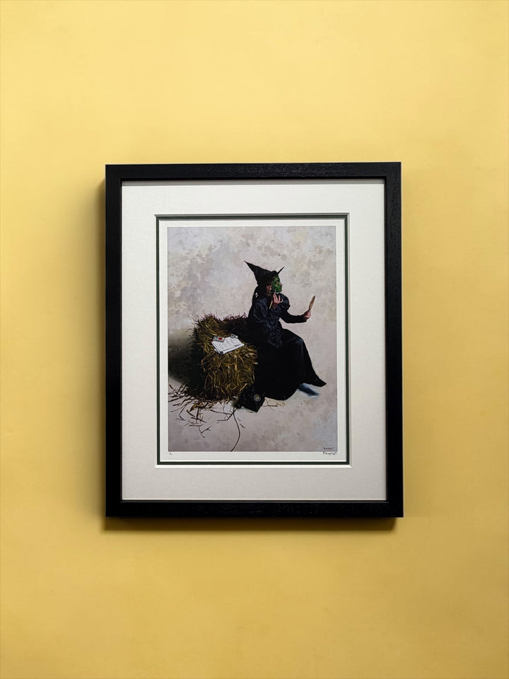 A framed art print depicting a witch in a black dress sitting on a nest of twigs, mounted on a solid yellow wall.