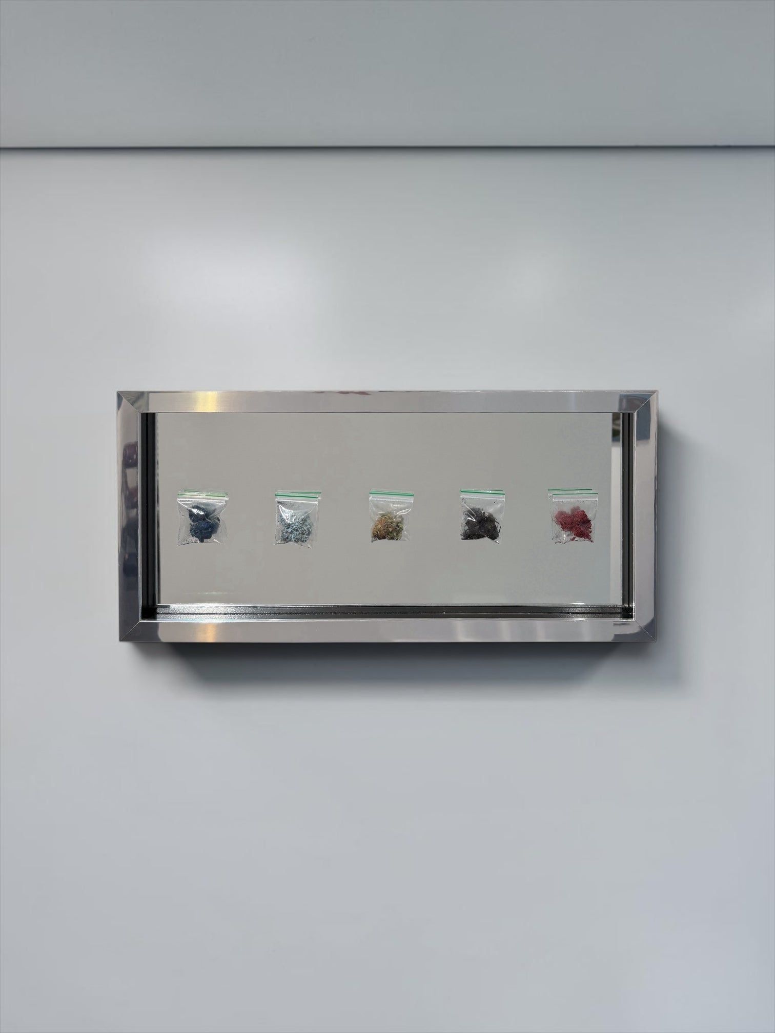 A rectangular stainless steel wall-mounted display case with a glass front, showing five small clear plastic bags containing different colored materials, mounted on a light gray wall.